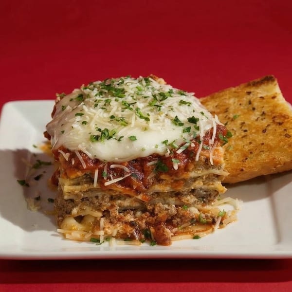 A tall slice of lasagna with melted cheese and parsley, served with garlic bread.