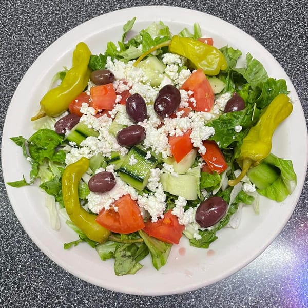 A white plate holds a vibrant Greek salad with lettuce, tomatoes, cucumbers, feta, olives, and pepperoncini.