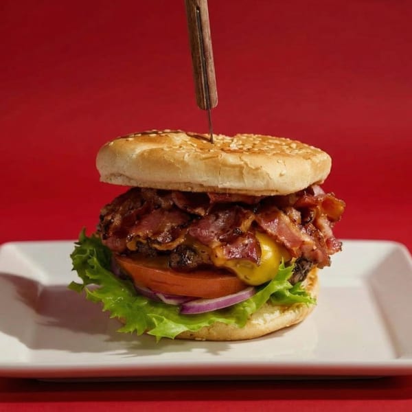 Bacon cheeseburger with lettuce, tomato, and onion, held together by a knife on a white plate.